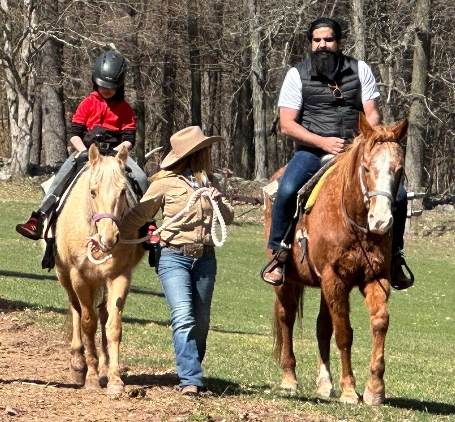 A child in a riding helmet on a led pony, with a handler on foot and a second rider alongside, on a wooded trail