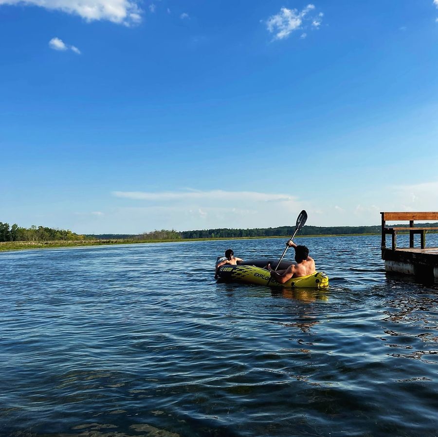 Two people paddling a yellow inflatable kayak on a lake beside a wooden dock, photographed from behind