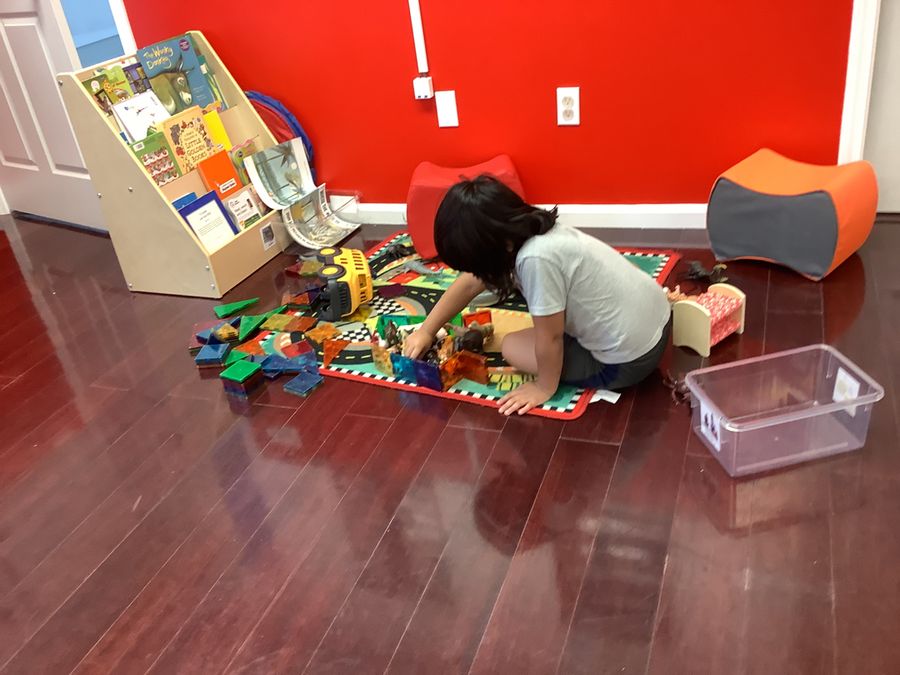A child playing with magnetic tiles and toy vehicles on a play mat beside a book display and sensory seating
